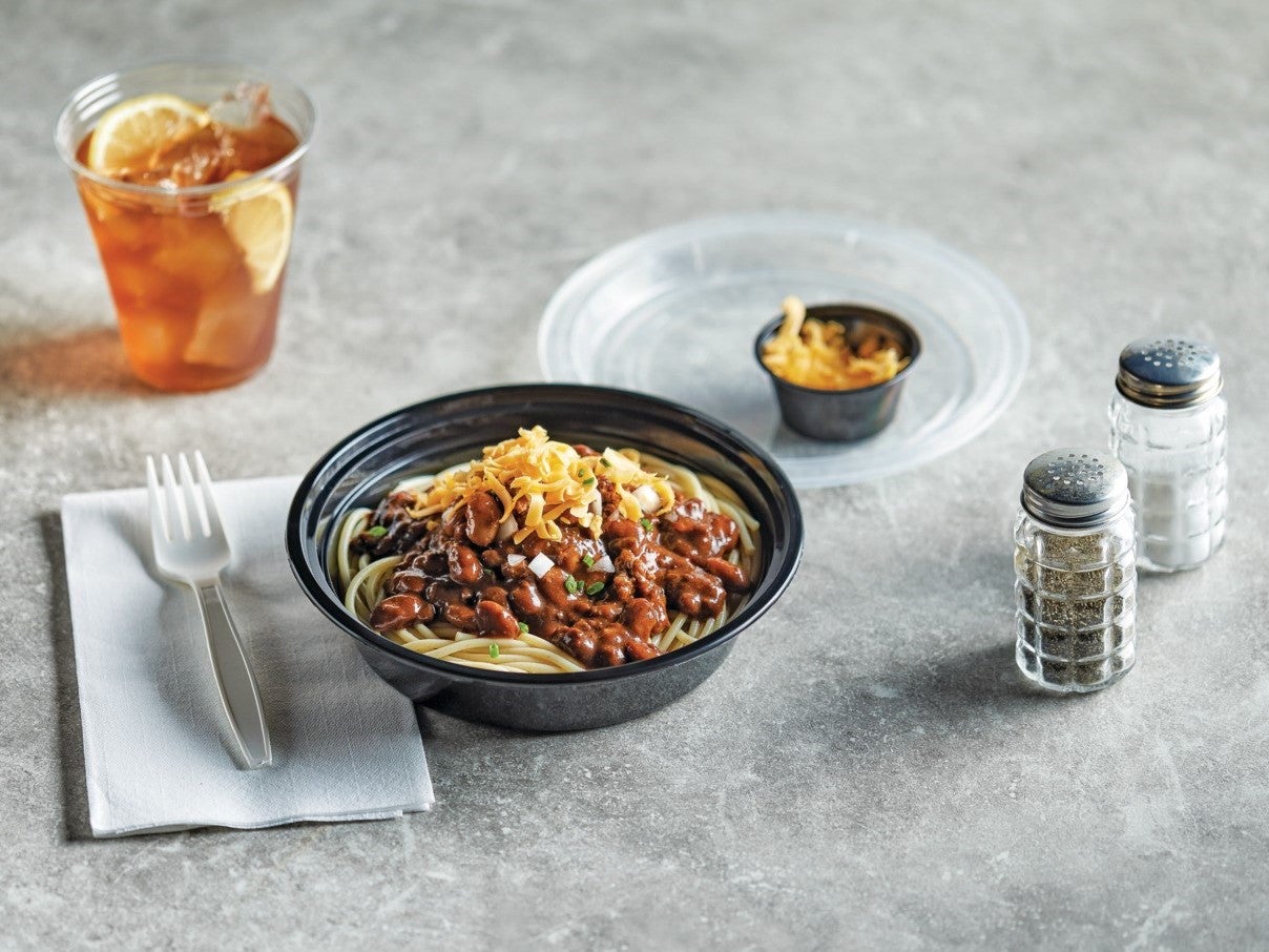 Un plato negro con pasta y fríjoles con chili, acompañado de cubiertos y bebida