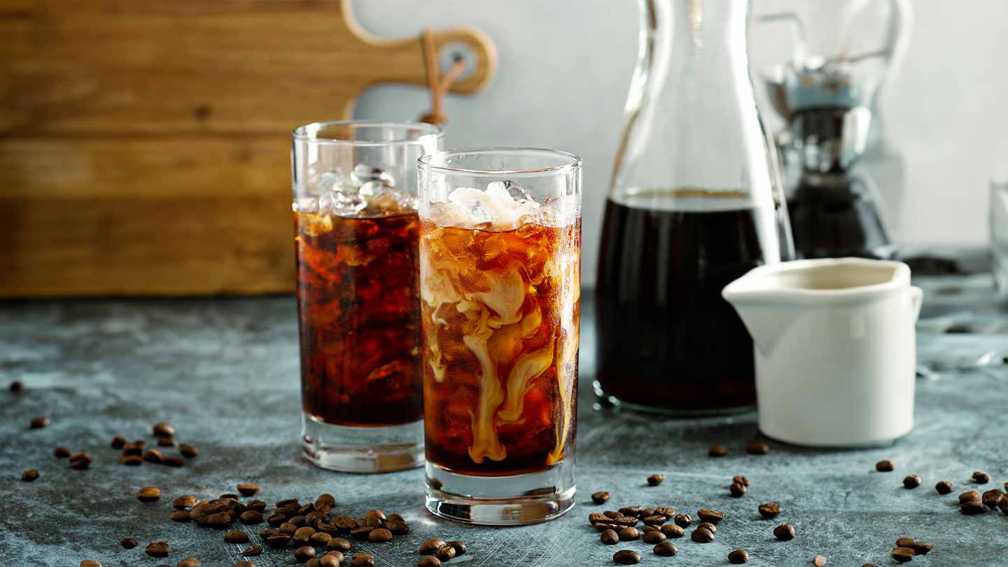 Dos vasos de café frío con hielo y leche sobre una mesa junto con granos de café tostados y una jarra con la bebida