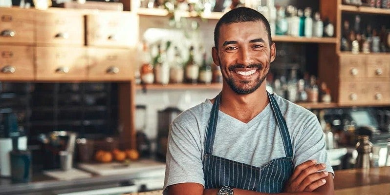 Un barista cruzado de brazos sonríe y a su espalda está la cocina de una cafetería equipada con ingredientes e instrumentos