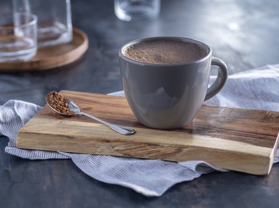 Una taza gris de chocolate caliente para máquinas, sobre una tabla de madera, con una cucharada de cacao en polvo.
