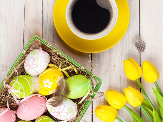 Sobre una mesa de madera 5 rosas amarillas, una taza de café y una canasta con dulces de pascua como huevos decorados
