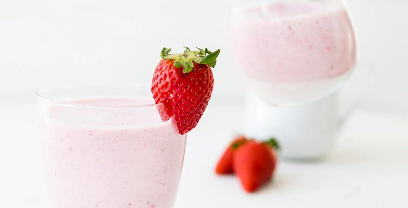 Batido en un vaso y en el borde una fresa anclada como elemento decorativo, al fondo una taza blanca