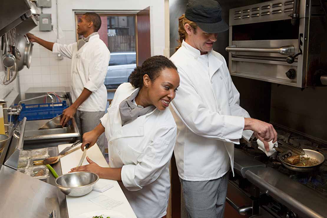 En una cocina industrial, una chef observa cómo otro fríe una proteína mientras un chef más lava las ollas