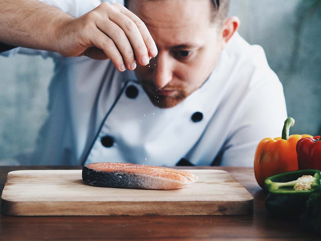 Chef sazona cuidadosamente una rodaja de pescado crudo sobre una tabla de picar junto a unos pimientos