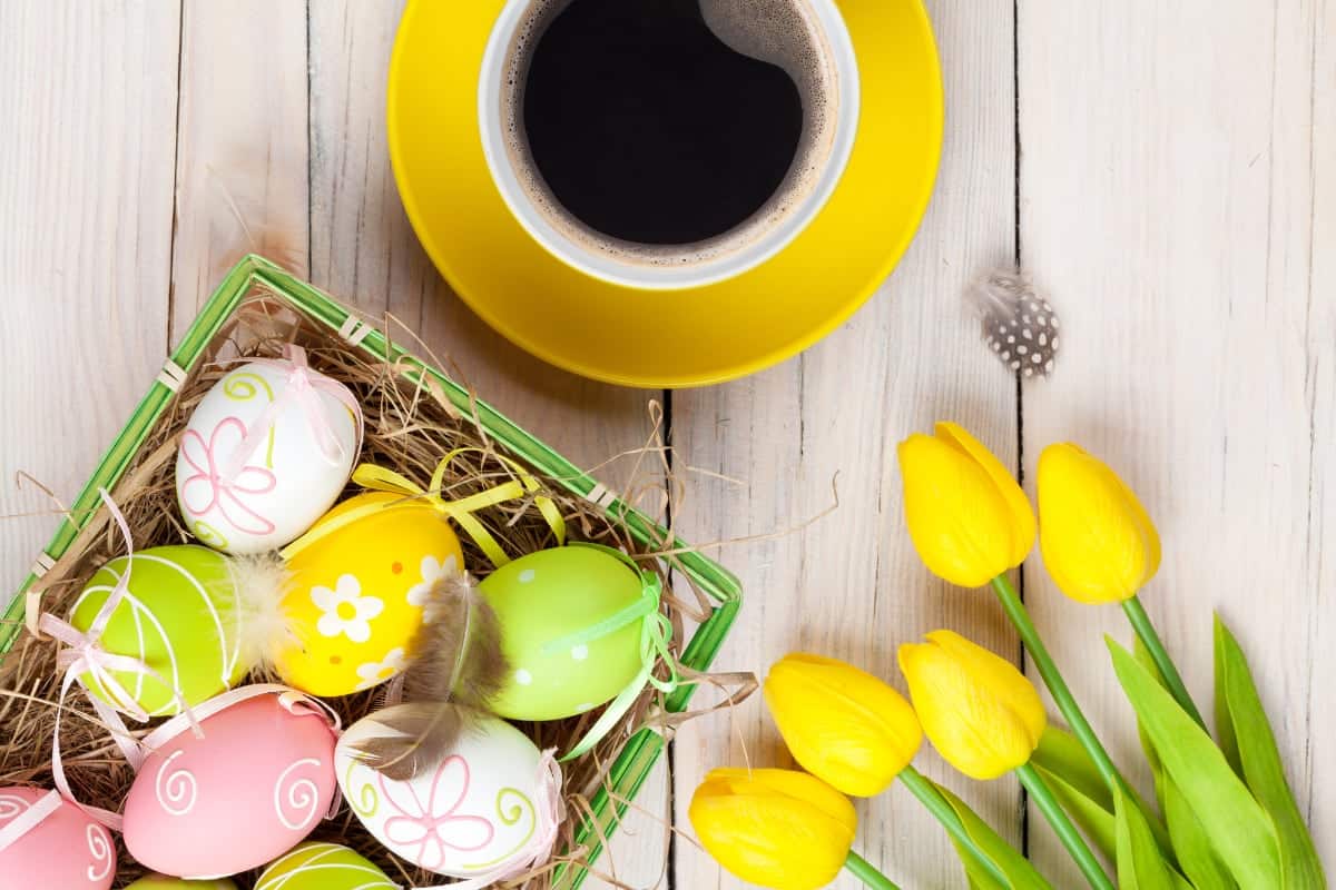 Sobre una mesa de madera 5 rosas amarillas, una taza de café y una canasta con dulces de pascua como huevos decorados