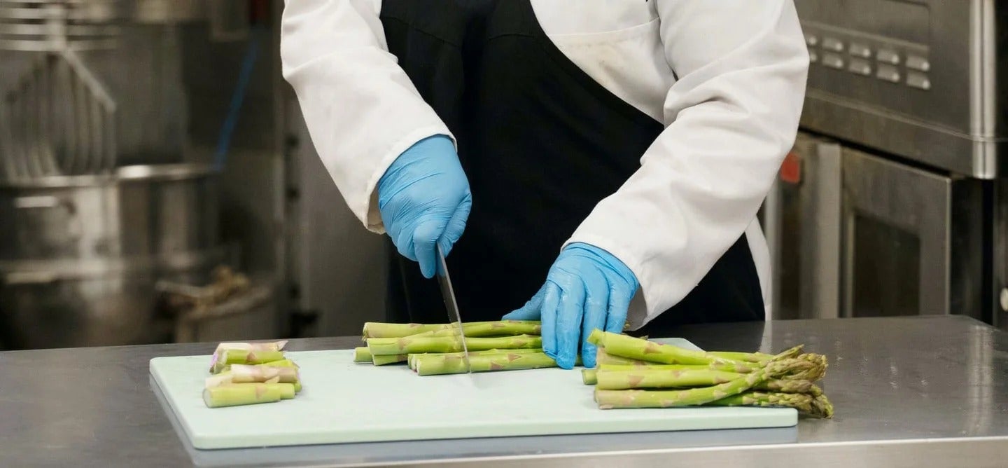 Chef con guantes azules cortando espárragos sobre tabla blanca