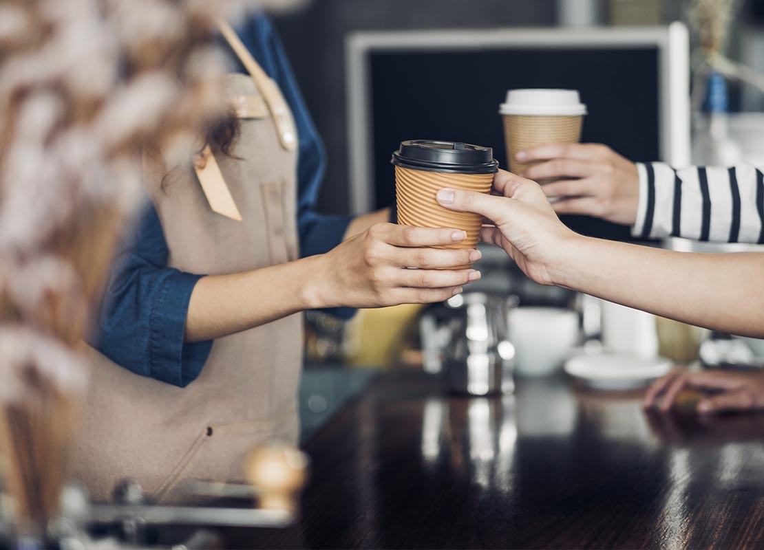 Empleada entrega por medio de take away a sus clientes vasos de cafés tapados en la barra de una cafetería