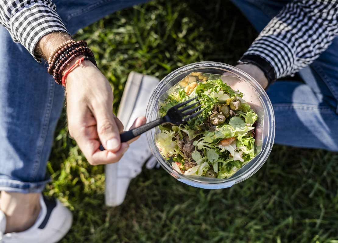 Hombre sentado en el césped sostiene tenedor y ensalada, la cual pidió por un servicio take away