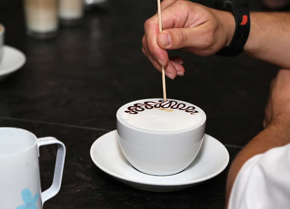 Sobre una mesa, mano de barista usa un palillo para un diseño de latte art con la técnica etching