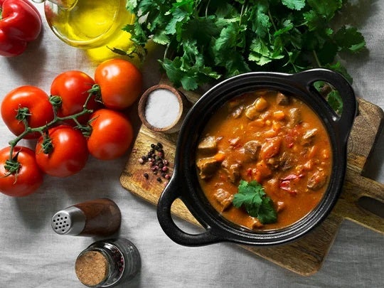 Olla de estofado rodeada por tomates, aceite, hierbas aromáticas y otros alimentos o productos culinarios