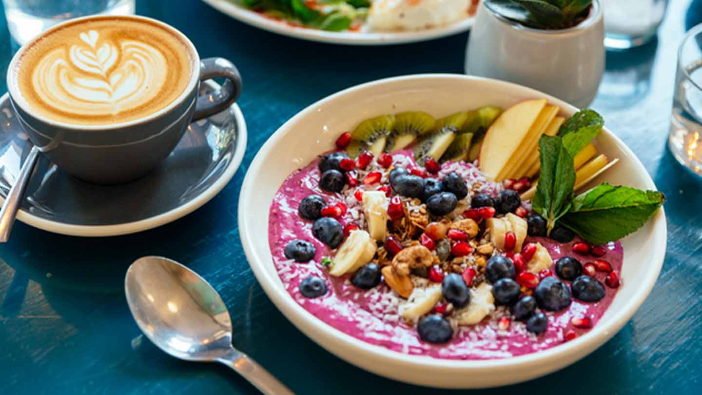 Servido a la mesa un bowl con yogurt y fruta, cappuccino y vasos de agua, que componen un desayuno
