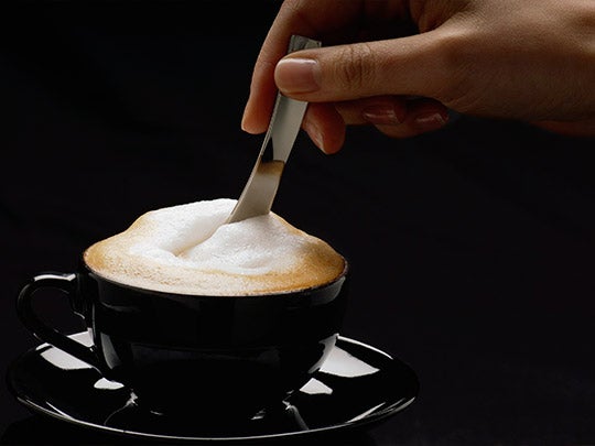 Mano de una mujer mezcla con una cuchara un cappuccino NESCAFÉ® servido en una taza negra