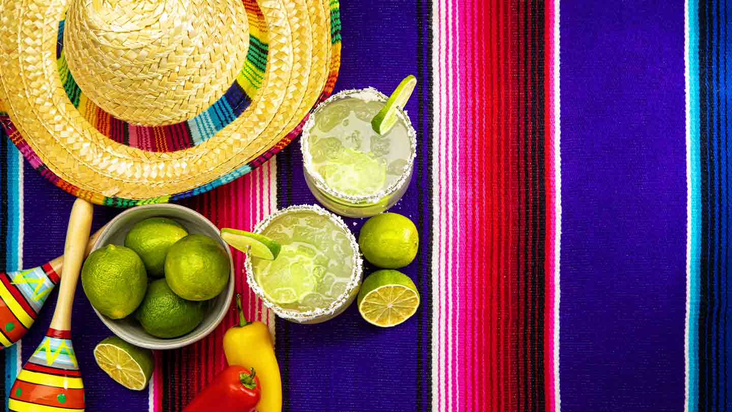 Dos vasos de michelada con rodajas de limón en el borde y al lado la fruta, un sombrero y maracas