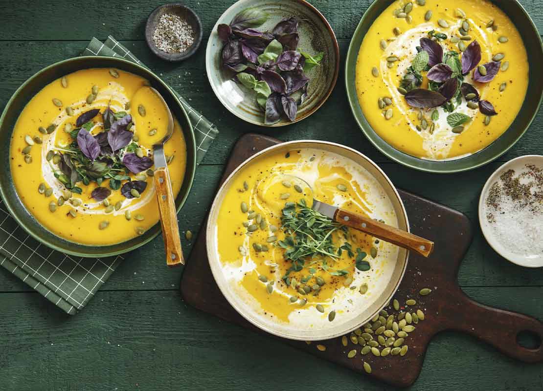 Tres platos de crema de zapallo decorados con semillas y hojas de albahaca morada y verde