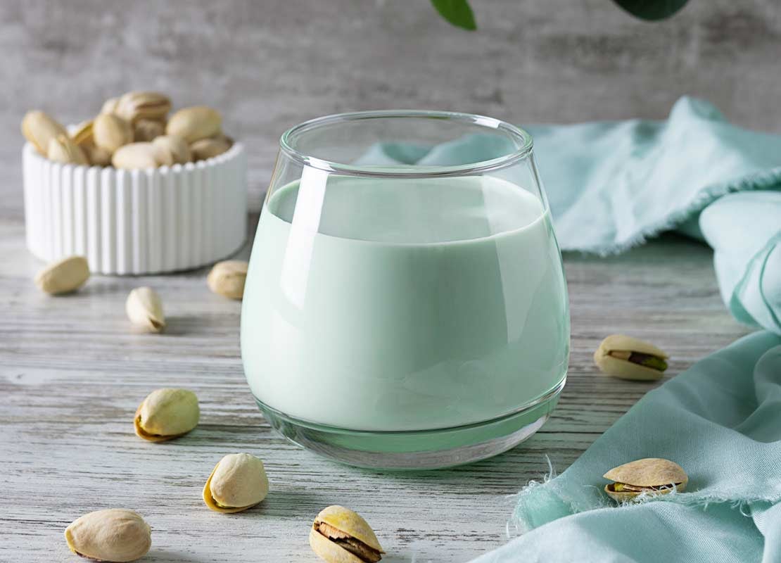 Bebida vegetal de pistacho en un vaso de cristal rodeada de este fruto seco sobre una mesa de madera