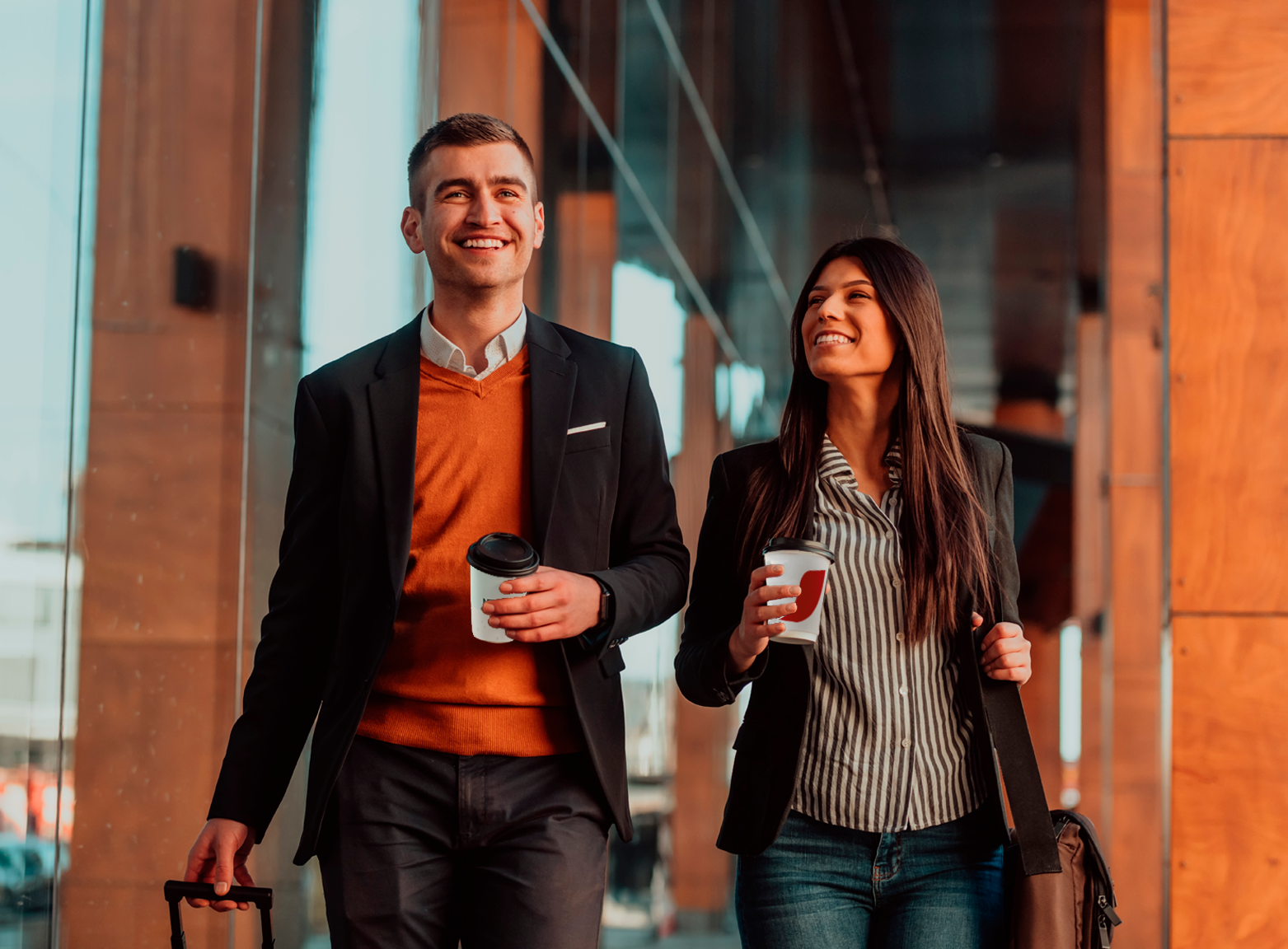 Hombre y mujer sonrientes, caminando en aeropuerto con bebidas de NESCAFÉ en mano y equipaje