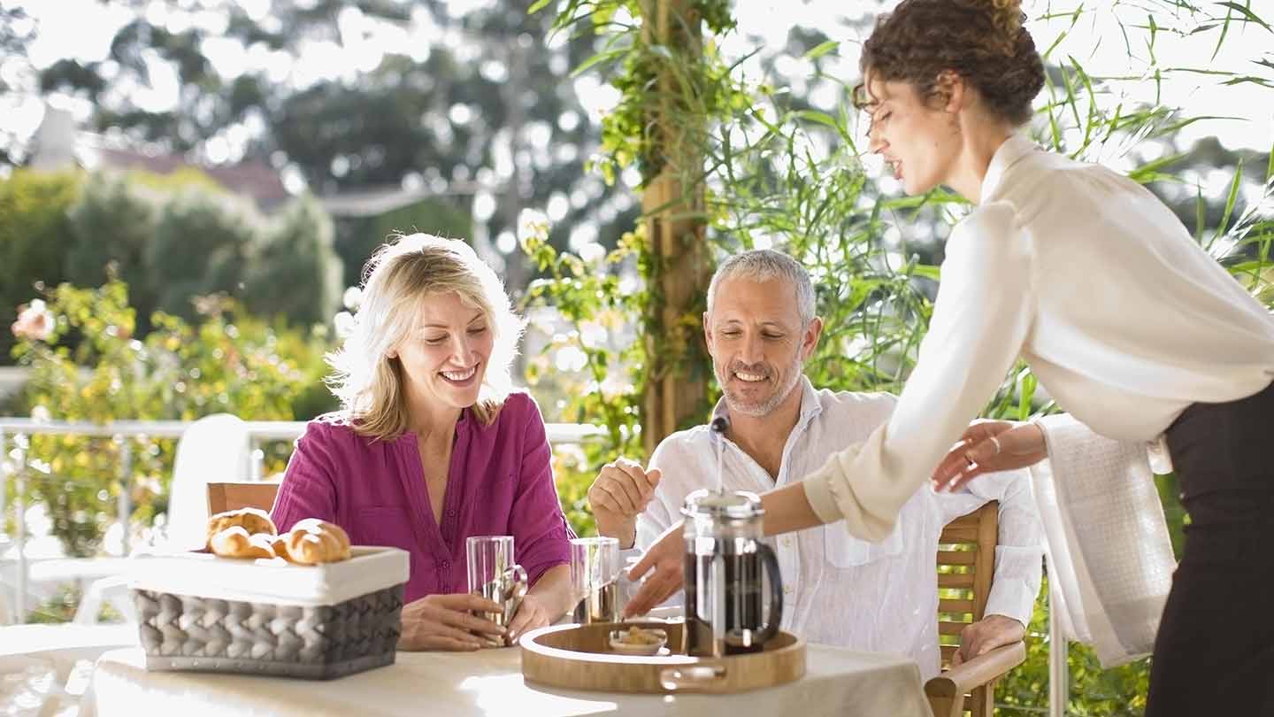 Mesera entrega el desayuno a la mesa de una pareja de clientes en un restaurante al aire libre