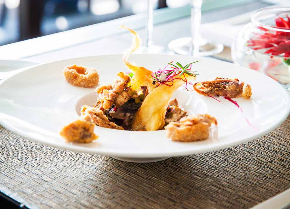 Comida de autor servida en un plato hondo de manera sofisticada, al fondo copas de cristal