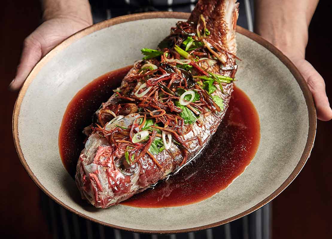 Chef sostiene un plato con pescado entero cocido al vapor, servido con una salsa oscura, chile y cebollín