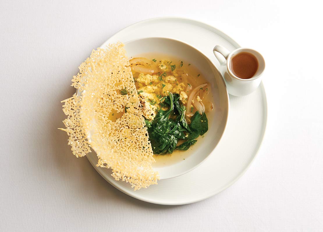 Plato de Stracciatella (sopa) que combina huevo, queso y hojas verdes, decorado con tuile en el borde y salsera
