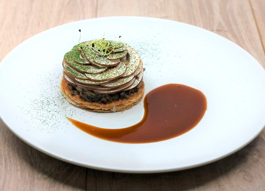 Plato de tarta fina con verduras y láminas de champiñones apiladas tipo teja y decorado con una salsa espesa