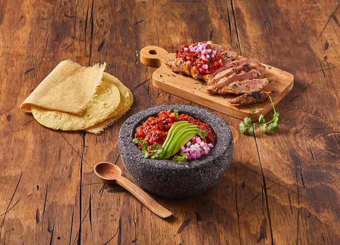 Molcajete con salsa roja, aguacate y cebolla, acompañado de tortillas y carne asada en rodajas sobre tabla