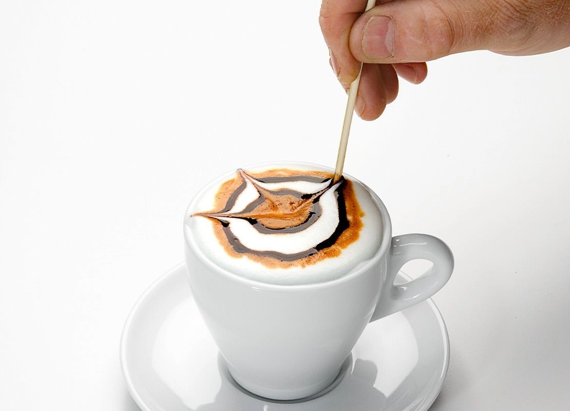 Mano decorando un café con arte latte usando un palillo sobre espuma y jarabe