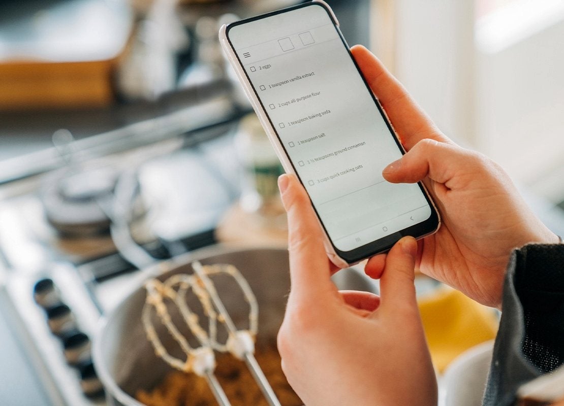 Manos de mujer sostienen un celular que muestra una lista de ingredientes, al fondo utensilios de cocina
