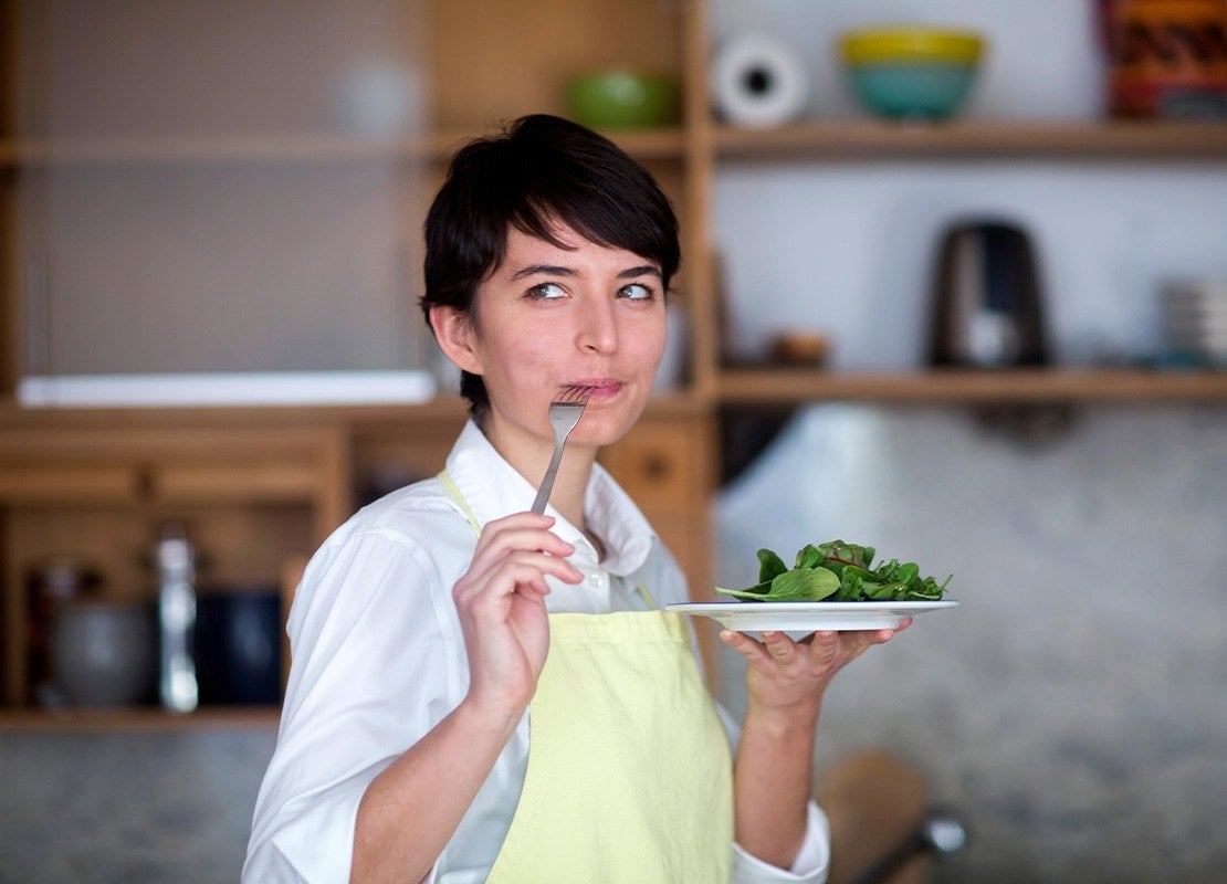 En una cocina, una chef sostiene un tenedor en su boca mientras en la otra mano tiene un plato de ensalada