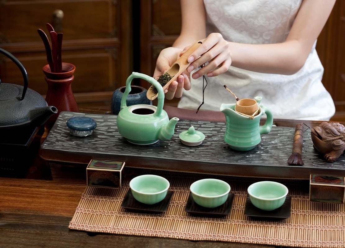 Mujer vierte hojas en una tetera de porcelana sobre una mesa y rodeada de otros utensilios para elaborar té