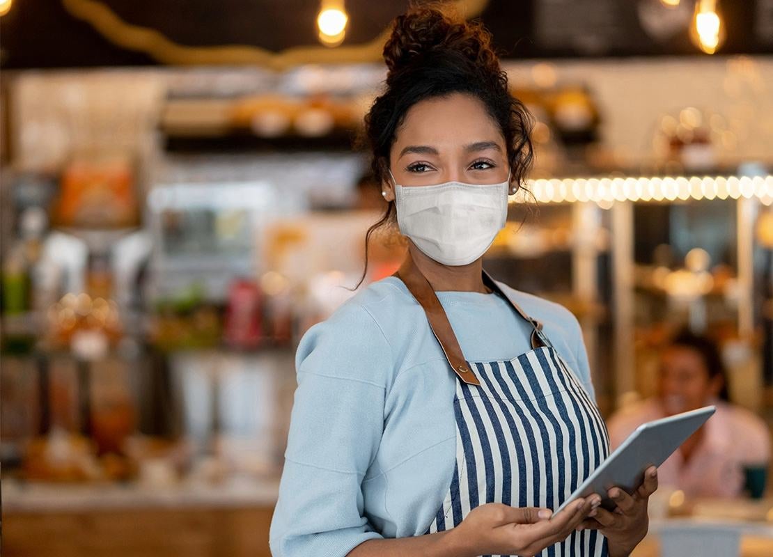 Mesera con mascarilla y delantal sostiene una tablet en una cafetería, lista para tomar órdenes