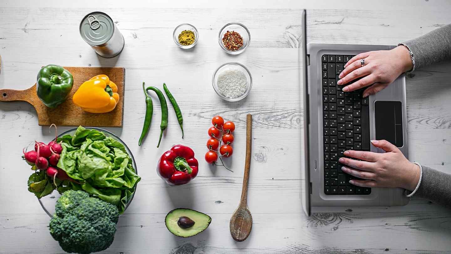 Ingredientes frescos como vegetales, especias y arroz frente a una laptop usada por las manos de una mujer