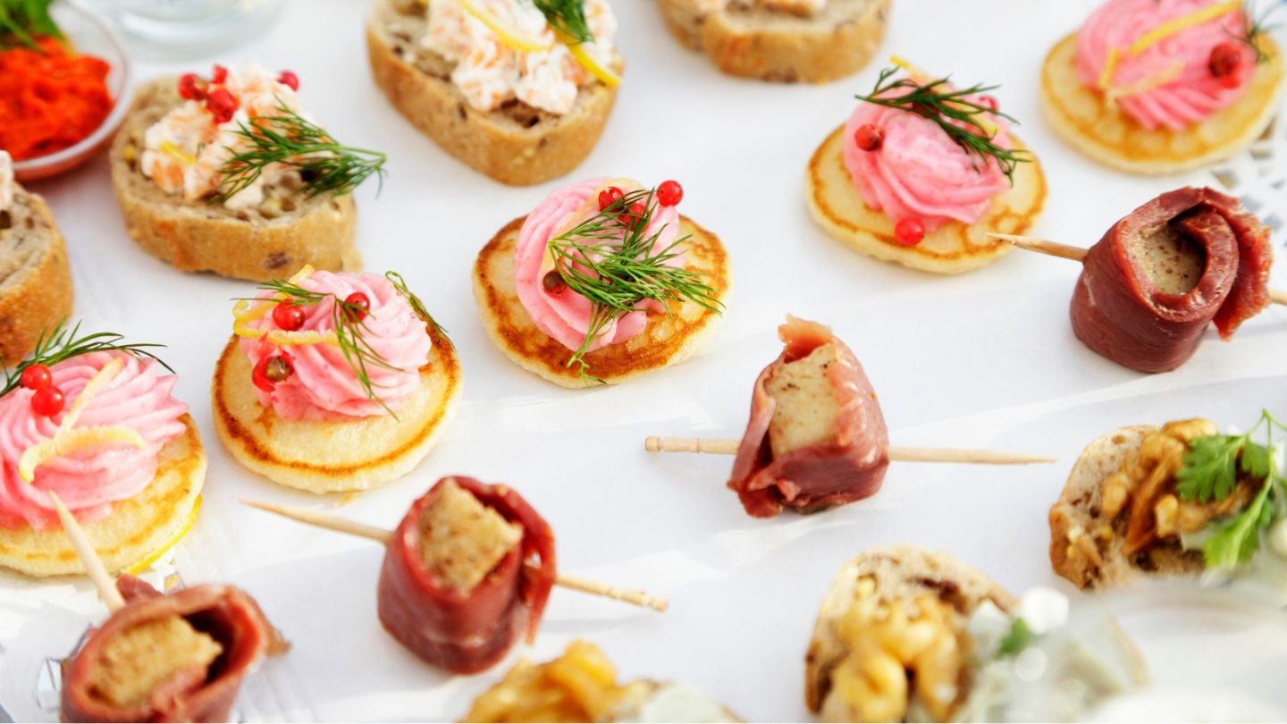 Variedad de canapés decorados con crema, eneldo, frutos rojos y carnes frías, dispuestos sobre una bandeja