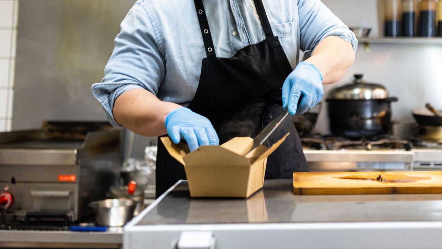 Chef con guantes y delantal empacando comida en una caja para llevar dentro de una dark kitchen profesional