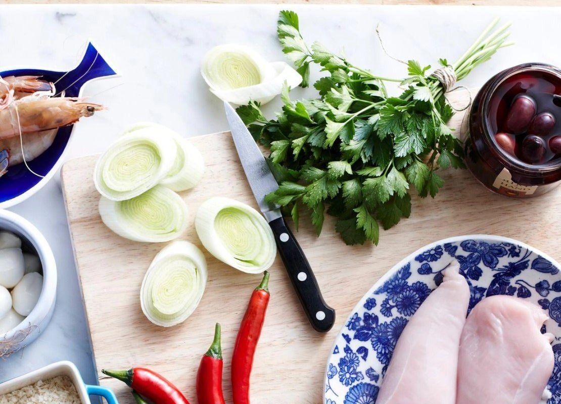 Ingredientes frescos como pollo, verduras y camarones junto a un cuchillo sobre una tabla.