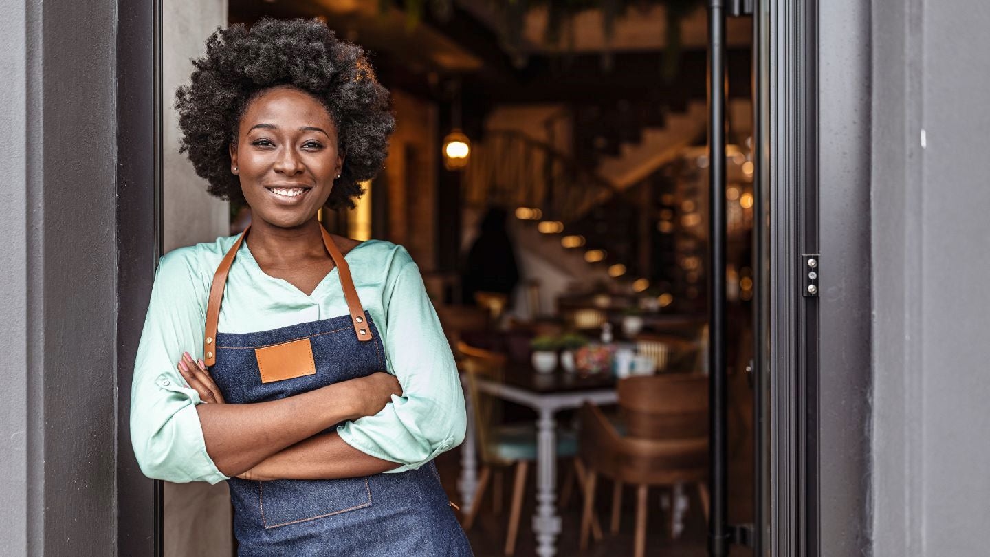 Dueña de negocio gastronómico con delantal frente a la puerta de su establecimiento