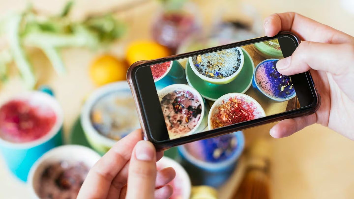 Persona tomando una foto con el celular a varias tazas coloridas de bebidas decoradas con flores y semillas