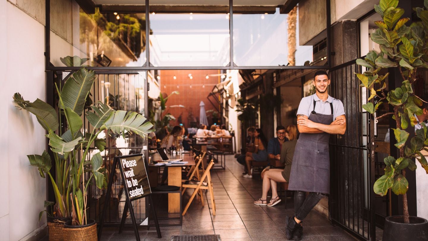 Entrada de un restaurante con plantas y mesas al aire libre, creando un ambiente tipo jardín gastronómico.