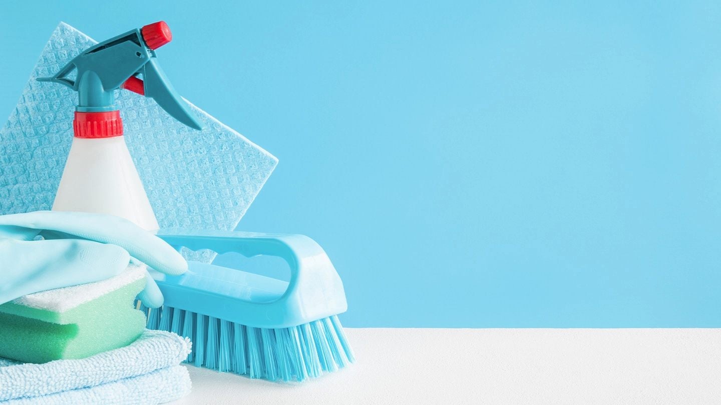 Artículos de limpieza como guantes, esponjas, cepillo, trapo y atomizador sobre fondo azul.