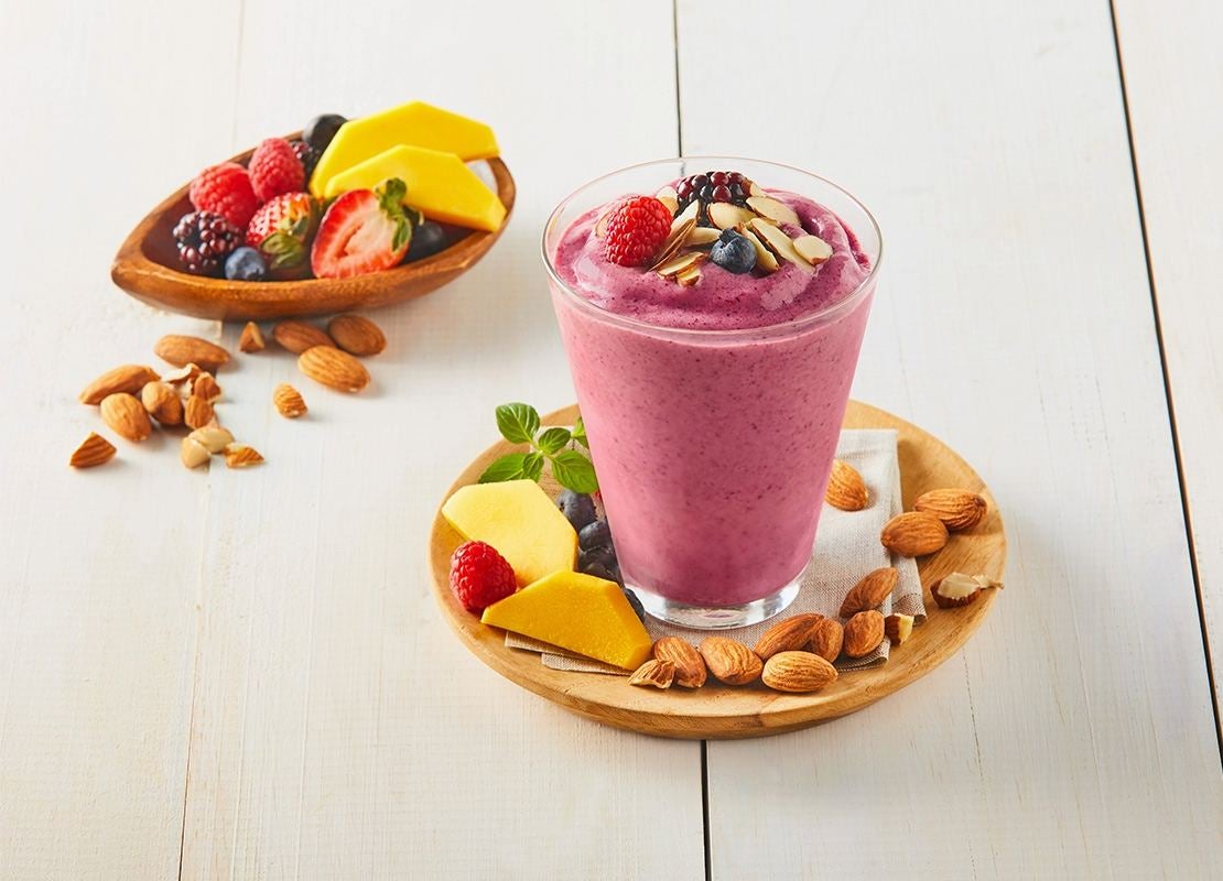 Vaso de kombucha berries decorado con mango, frutos rojos y almendras, al fondo ingredientes frescos