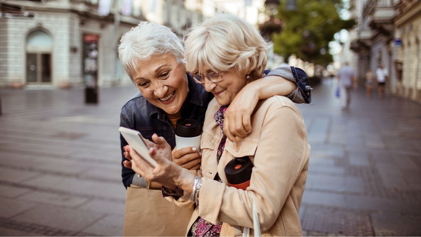 Dos mujeres mayores sonrientes observan un celular mientras pasean por una ciudad con café en mano