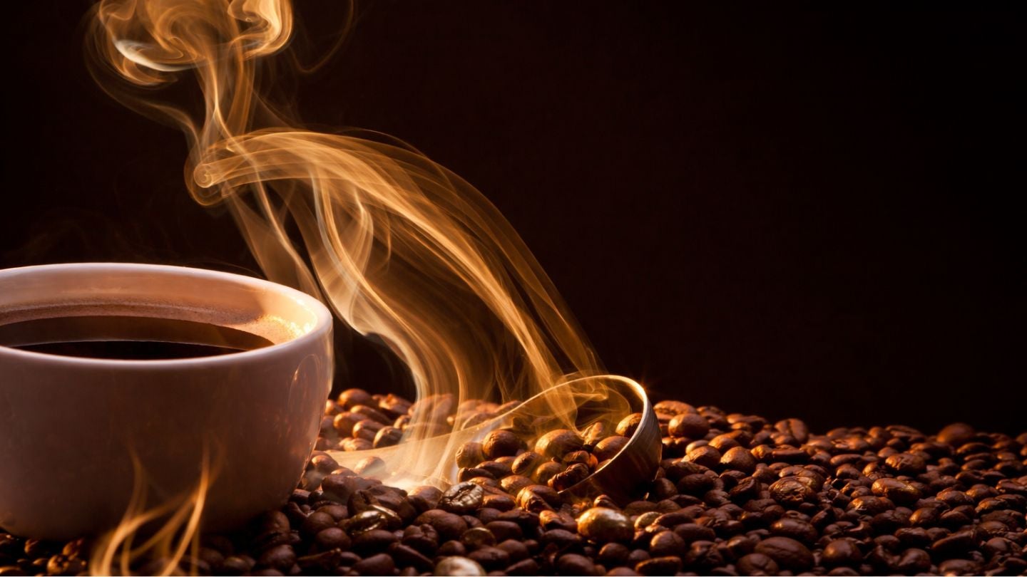 Taza de café humeante entre granos tostados, con el vapor formando curvas en un ambiente oscuro