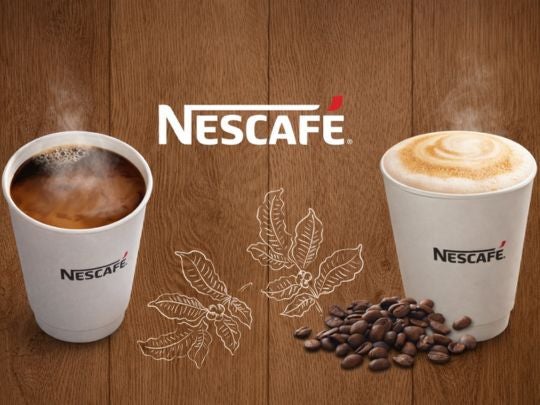 Dos vasos de NESCAFÉ con café americano y cappuccino de calidad, al pie, granos tostados