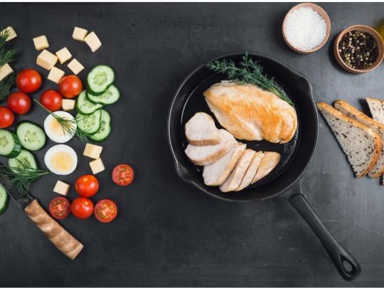 Sartén con pollo rodeado de verduras frescas, pan y especias sobre una mesa negra