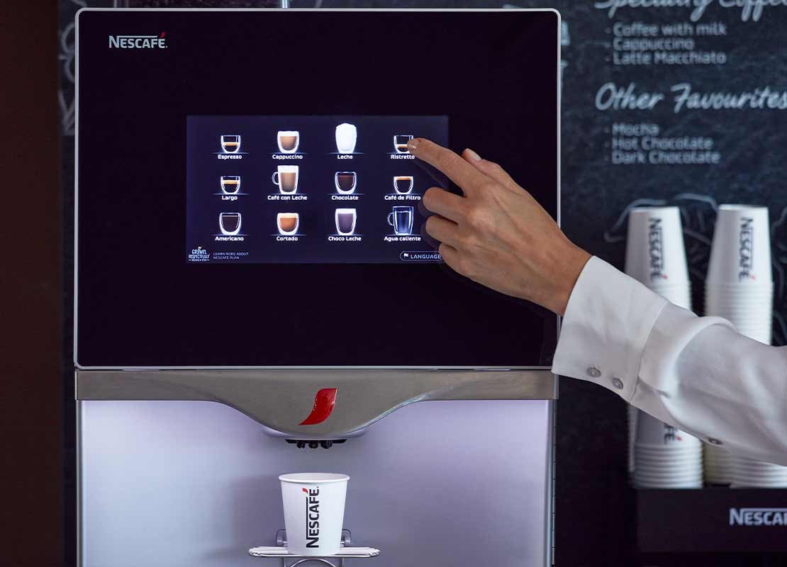 Una máquina NESCAFÉ® dispensa una bebida mientras un usuario selecciona opciones en la pantalla táctil.