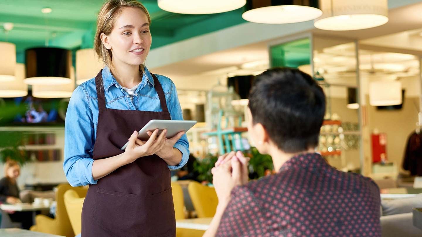 Mesera atiende a un cliente en restaurante, escuchando su solicitud con atención y profesionalismo.