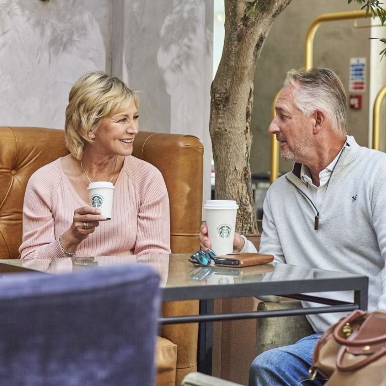 Pareja de adultos mayores conversando en lounge de hotel mientras disfrutan café caliente.