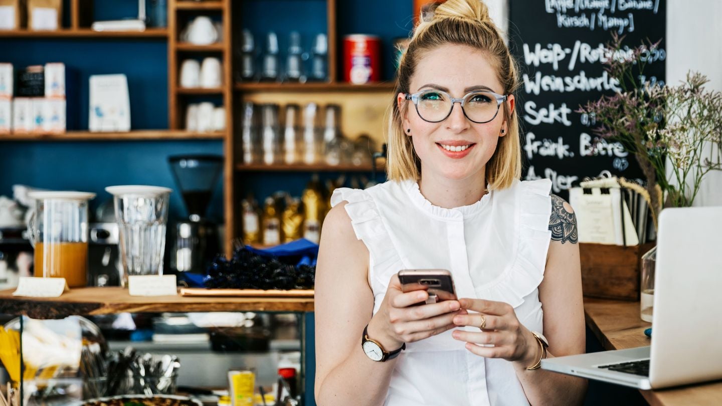 Mujer en cafetería usando celular y laptop, gestionando tareas en entorno de negocio.