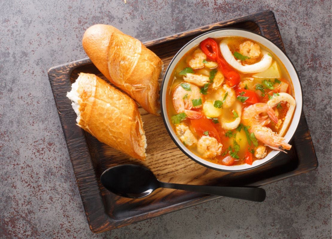 Sopa con mariscos y vegetales servida en tazón con pan sobre bandeja de madera