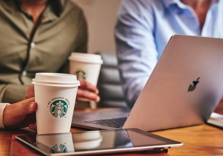Personas en reunión de oficina compartiendo café para llevar mientras trabajan con laptops.